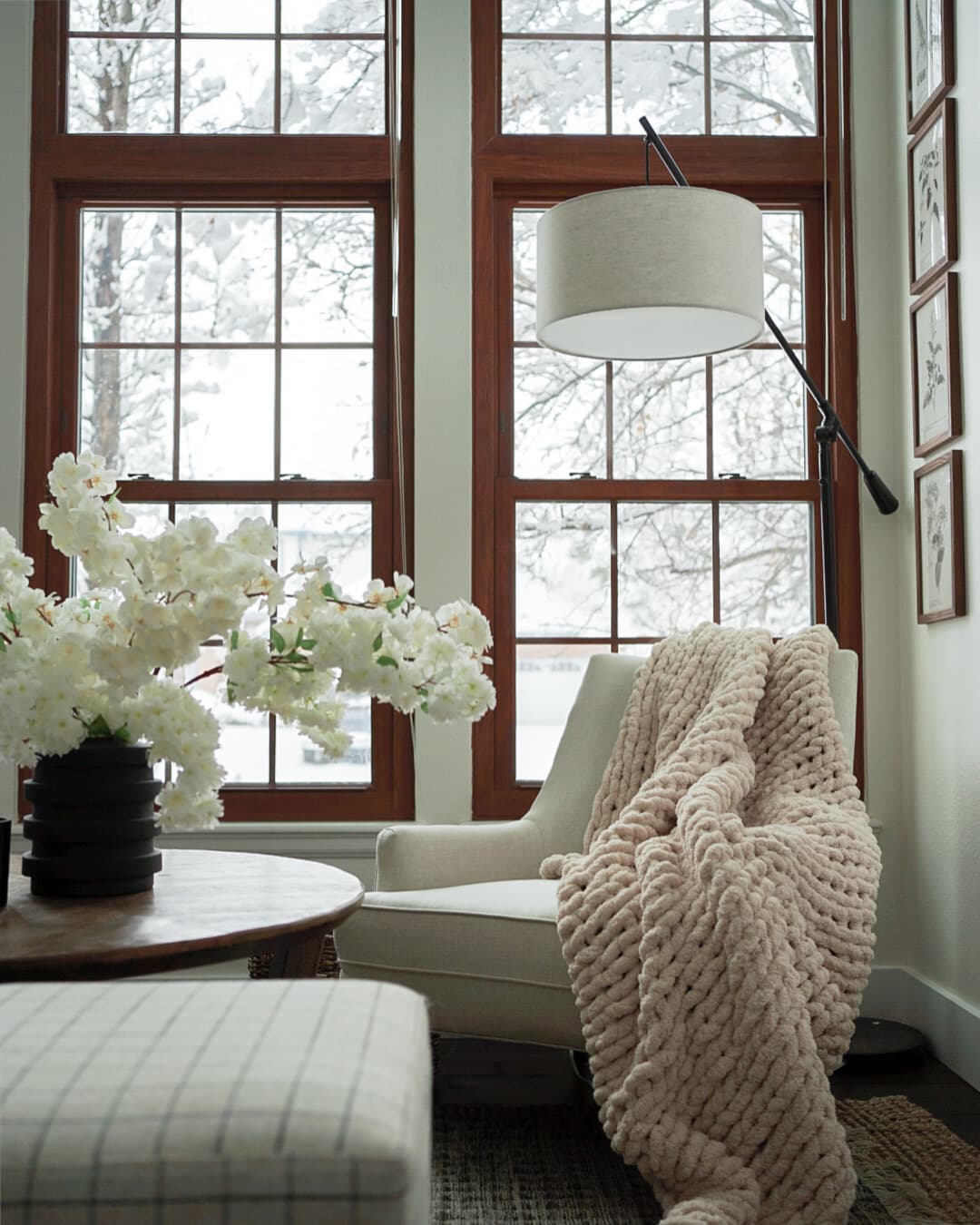 Reading nook by tall windows with a cream armchair, plaid ottoman, and a knit throw in a Highlands Ranch home