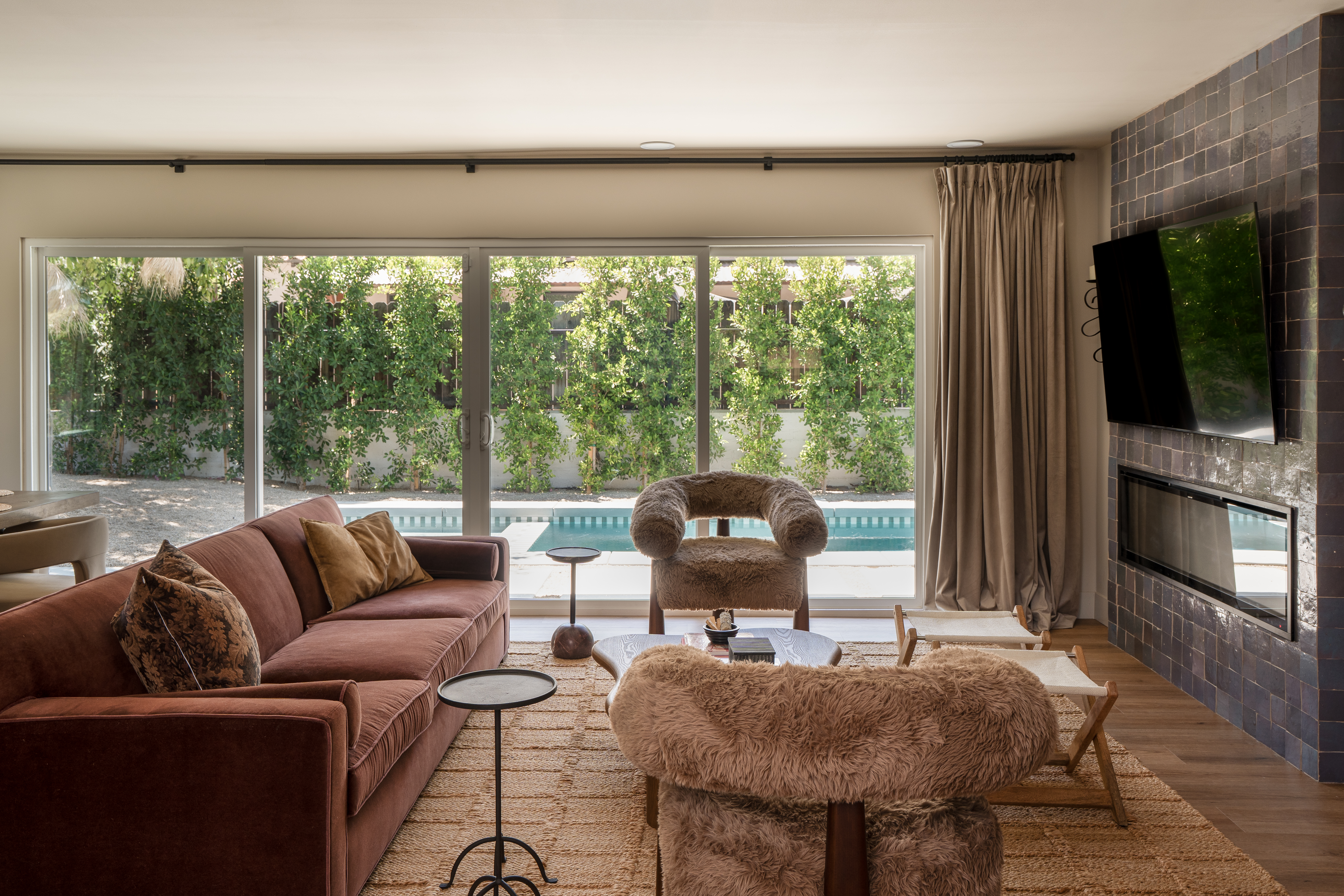 Palm Springs living room with rust velvet sofa, shearling armchairs, and a deep blue glazed tile fireplace wall, opening through sliding glass doors to the pool, designed by Halcyon Haus